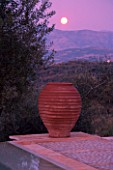DUSK VIEW ACROSS SWIMMING POOL TOWARDS TERRACOTTA URN WITH FULL MOON  IN GINA PRICES CORFU GARDEN.