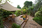 EXOTIC SHADE PLANTING OF GRASSES  PHORMIUM  BAMBOO  ROSEMARY AND OTHER FOLIAGE PLANTS WITH CANOPY OVER DECKED DINING AREA IN SMALL TOWN GRAVEL GARDEN BY KATHY TAYLOR  LONDON.