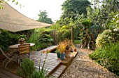 EXOTIC SHADE PLANTING OF GRASSES  PHORMIUM  BAMBOO  ROSEMARY AND OTHER FOLIAGE PLANTS WITH CANOPY OVER DECKED DINING AREA IN SMALL TOWN GRAVEL GARDEN BY KATHY TAYLOR  LONDON.