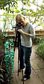 BLOND HAIRED GIRL WITH 35MM SLR DIGITAL CAMERA ON A TRIPOD IN A GREENHOUSE TAKING PHOTOGRAPHS OF A JADE VINE