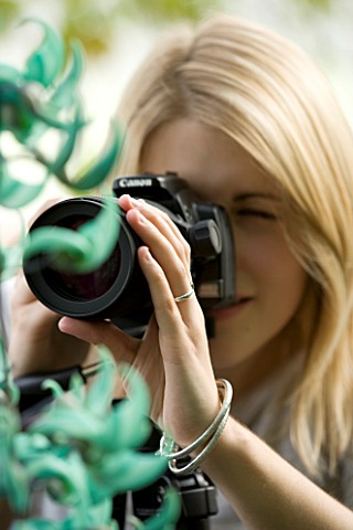 BLOND_HAIRED_GIRL_WITH_35MM_SLR_DIGITAL_CAMERA_ON_A_TRIPOD_IN_A_GREENHOUSE_TAKING_PHOTOGRAPHS_OF_A_J