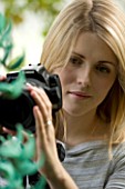 BLOND HAIRED GIRL WITH 35MM SLR DIGITAL CAMERA ON A TRIPOD IN A GREENHOUSE TAKING PHOTOGRAPHS OF A JADE VINE