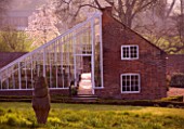 KELMARSH HALL  NORTHAMPTONSHIRE: THE GREENHOUSE IN THE WALLED GARDEN AT DUSK IN THE SPRING WITH WOODEN SCULPTURE IN FOREGROUND