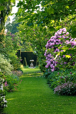 WARDINGTON_MANOR_GARDEN__OXFORDSHIRE_GRASS_PATH_LEADS_TO_AN_URN_PAST_RHODODENDRONS_AND_AZALEAS_IN_SP