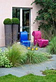 GARDEN DESIGNED BY CHARLOTTE SANDERSON: GREY SLATE PATH LEADS TO PATIO WITH PINK AND BLUE MODERN CHAIRS AND TWO TALL CYCLINDRICAL CONTAINERS PLANTED WITH BOX