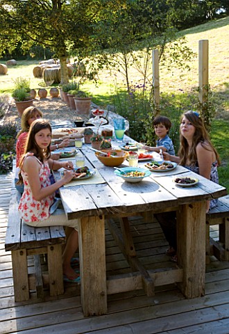CLARE_MATTHEWS_GARDEN__DEVON_CLARE_AND_FAMILY_SIT_DOWN_TO_AN_AL_FRESCO_LUNCH_AT_THE_TABLE_OUTDOOR_DI
