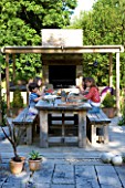 CLARE MATTHEWS GARDEN  DEVON. CLARE AND FAMILY SIT DOWN TO AN AL FRESCO LUNCH AT THE OUTDOOR DINING TABLE. PERGOLA  OUTDOOR OVEN. DINING.  DESIGNER CLARE MATTHEWS