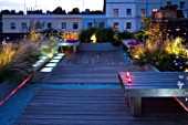 ROOF GARDEN HOLLAND PARK  LONDON.DESIGNER: CHARLOTTE ROWE. DECKED TERRACE IN EVENING WITH PINK & WHITE LED LIGHTING & BLUE GLASS GRAVEL. CANDLES ON WOODEN BENCHON BENCH