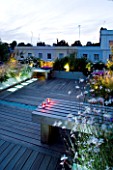 ROOF GARDEN  HOLLAND PARK  LONDON. DESIGNER: CHARLOTTE ROWE. DECKED TERRACE IN EVENING WITH LED LIGHTING  CANDLES ON WOODEN BENCH  STIPA TENUISSIMA  GAURA LINDHEIMERI