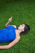 TEENAGE GIRL (16-17 YEARS) IN BLUE TOP LYING ON GRASS WRITING LISTENING TO MP3 PLAYER. MUSIC  TEENAGE GIRLS  ONE TEENAGE GIRL ONLY  CASUAL CLOTHING  STUDENT  COLLEGE  IN THE PARK