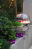 DESIGNER - CHARLOTTE ROWE  LONDON: CHARLOTTE ROWES OWN GARDEN AT NIGHT - RENDERED RAISED BED BESIDE FIREPLACE WITH LIRIOPE MUSCARI  CANDLES AND A LARGE GLASS BOWL WITH GERBERAS