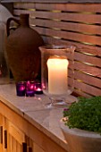 DESIGNER - CHARLOTTE ROWE  LONDON: CHARLOTTE ROWES OWN GARDEN AT NIGHT - CANDLES ON MARBLE SHELF  OLD TERRACOTTA URN AND WOODEN TRELLIS