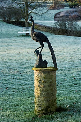 CERNEY_HOUSE_GARDEN__GLOUCESTERSHIREPEACOCK_SCULPTURE_ON_A_PEDESTAL_ON_THE_LAWN_IN_FONT_OF_THE_HOUSE