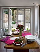 DESIGNER: CHARLOTTE ROWE  LONDON: THE DINING ROOM WITH TABLE WITH RED TULIPS IN A GLASS VASE  PINK NAPKINS AND FRESH STRAWBERRIES ON A PLATE. BEHIND IS THE GARDEN WITH OUTDOOR FIRE