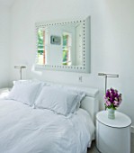 TANIA LAURIE  LONDON. WHITE BEDROOM WITH MIRROR ABOVE BED AND BEDSIDE TABLE WITH VASE OF SCENTED STOCK