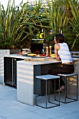 CONTEMPORARY FORMAL ROOF TERRACE/ GARDEN DESIGNED BY DATA NATURE ASSOCIATES: GIRL SITTING AT A TABLE BESIDE THE BARBEQUE