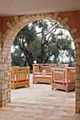 DESIGNER GINA PRICE - CORFU - VILLA ONEIRO - VIEW THROUGH ARCHWAY TO OUTDOOR PATIO WITH WOODEN SEATS AND CUSHIONS BESIDE THE VILLA