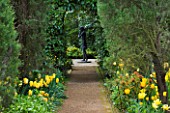PASHLEY MANOR GARDEN  EAST SUSSEX  SPRING :PATH THROUGH WOODLAND WITH YELLOW TULIPS AND VIEW THROUGH ARCH TO SCULPTURE  LITTLE FLAUTIST BY MARY COX