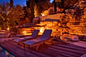 THE ROU ESTATE  CORFU  GREECE: DESIGNER: DOMINIC SKINNER - THE SWIMMING POOL AREA  LIT UP AT NIGHT WITH SUN LOUNGERS AND WATERFALL. LIGHTING