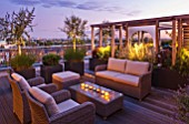 DESIGNER: CHARLOTTE ROWE  LONDON: ROOF GARDEN - A PLACE TO SIT - WICKER FURNITURE  WOODEN PERGOLA  CONTAINERS PLANTED WITH OLIVE TREES  PENNISETUM HAMELYN    PINUS MUGO. LIGHTING,  DECKS, DECKING, SCREENING