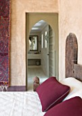DESIGNERS ERIC OSSART AND ARNAUD MAURIERES  MOROCCO: AL HOSSOUN - BEDROOM WITH WOODN BED  MAGENTA CUSHIONS AND CARPET ON WALL - VIEW THROUGH DOORWAY TO SHOWER ROOM
