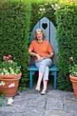 SANDHILL FARM HOUSE  HAMPSHIRE - DESIGNER ROSEMARY ALEXANDER SITTING IN A BLUE WOODEN SEAT SET INTO A YEW HEDGE AT THE END OF A PATH