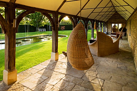 WHATLEY_MANOR__WILTSHIRE_SWING_SEAT_CHAIR_IN_THE_COTSWOLD_STONE_LOGGIA_BUILDING_WITH_LAWN__POOL_AND_