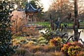 RHS GARDEN WISLEY  SURREY:  EVENING VIEW ACROSS THE LAKE AT SEVEN ACRES TO THE CHINESE PAGODA WITH HAMAMELIS APHRODITE  CAREX FLAGELLIFERA AND CAREX ASHIMENSIS EVERGOLD. WINTER