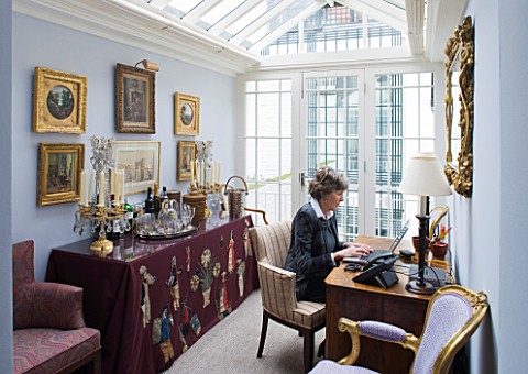 DESIGNER_JANE_CHURCHILL__JANE_CHURCHILL_SITS_AT_HER_DESK_IN_THE_CONSERVATORY_WITH_DRINKS_TABLE