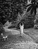 TREGOTHNAN  CORNWALL: BLACK AND WHITE IMAGE OF WHITE BENCH BENEATH RHODODENDRON RUSSELLIANUM - GIRL WALKING WITH TRUG