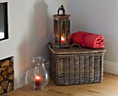 JACKY HOBBS HOUSE  LONDON: FIRESIDE SCENE WITH LOG PILE  WICKER BASKET RED THROW AND WOODEN AND GLASS STROM LANTERNS WITH CANDLES