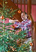 RICKYARD BARN  OXFORDSHIRE: CHRISTMAS - CLIVE NICHOLS ATTACHING BAUBLE TO THE CHRISTMAS TREE