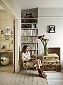 TWIG HUTCHINSON HOUSE  LONDON: TWIG IN WHITE LIVING ROOM WITH CHAIR  BOOKCASE  ALLIUMS IN GLASS JAR WITH SUITCASES BENEATH