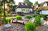 THE BOAT HOUSE: DESIGNER ARLETTE GARCIA - THE JAPANESE GARDEN WITH WOODEN BRIDGE  GRAVEL AND IRIS SIBIRICA - DECK WITH TABLE AND CHAIRS IN FRONT OF HOUSE
