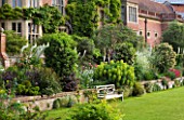 GLYNDEBOURNE, EAST SUSSEX: VIEW TO THE OPERA HOUSE OVER THE DOUBLE HERBACEOUS BORDERS WITH WHITE FOXTAIL LILIES - EREMERUS - AND EUPHORBIA - WOODEN BENCH / SEAT ON LAWN