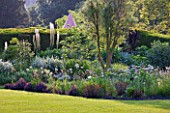 GLYNDEBOURNE, EAST SUSSEX: VIEW THROUGH YEW TREES TO LAWN AND BORDER WITH EREMURUS, ALLIUMS AND BERBERIS - BEYOND IS THE CHAMPAGNE TENT - HERBACEOUS, SUMMER