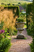 GLYNDEBOURNE, EAST SUSSEX: VIEW ALONG BRICK PATH TO STONE URN AND GAP THROUGH YEW HEDGES - STIPA GIGANTEA ON LEFT - FOCAL POINT, COUNTRY GARDEN
