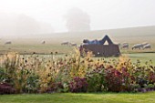 GLYNDEBOURNE, EAST SUSSEX: THE MAIN LAWN IN MIST WITH SCULPTURE IN BRONZE AND COR - TEN STEEL BY SEAN  HENRY ENTITLED CATAFALQUE, BORDER WITH SEDUMS AND GAURA LINDHEIMERI