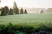 ROCKCLIFFE HOUSE, GLOUCESTERSHIRE: VIEW ACROSS HAHA TO BRONZE SCULPTURE SOUTHERN SHADE BY NIGEL HALL. MIST, FOG, GREEN, SUMMER - CLIPPED TOPIARY BEECH PYRAMIDS