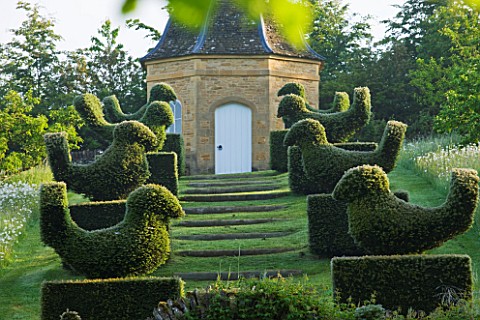 ROCKCLIFFE_HOUSE_GLOUCESTERSHIRE_ROW_OF_CLIPPED_TOPIARY_BIRDS_LEADING_UP_TO_DOVECOTE_SUMMER_COUNTRY_
