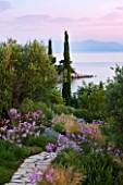 CORFU, GREECE - THE KASSIOPIA ESTATE: VIEW OUT TO SEA WITH STONE PATH, OLIVE AND CYPRESS TREE, STIPA TENUISSIMA AND TULBAGHIA VIOLACEA
