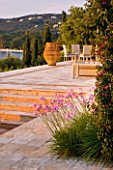 CORFU, GREECE - THE KASSIOPIA ESTATE: STONE TERRACE WITH SEATING AREA, HUGE STONE URN AND PINK FLOWERS OF TULBAGHIA VIOLACEA WITH BOUGAINVILLEA