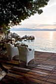 CORFU, GREECE - THE KASSIOPIA ESTATE: VIEW LOOKING OUT TO SEA AT SUNSET ON DECKED TERRACE WITH RATTAN CHAIRS. ALBANIAN MOUNTAINS IN THE DISTANCE