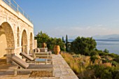 CORFU, GREECE - THE KASSIOPIA ESTATE. THE TERRACE WITH SUN LOUNGERS, TERRACOTTA URNS AND PLANTING BY JENNIFER GAY, LOOKING OUT TO SEA