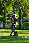 POULTON HOUSE GARDEN, WILTSHIRE: LAWN WITH IRISH YEWS AND ABSTRACT BRONZE STATUE / SCULPTURE CALLED CHAIN OF EVENTS BY TONY CRAGG