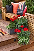 BEN DE LISI HOUSE AND GARDEN  LONDON@ A PLACE TO SIT - WOODEN BENCH WITH CUSHIONS AND SQUARE CONTAINERS PLANTED WITH RED GERANIUMS