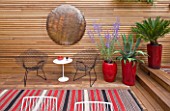 BEN DE LISI HOUSE AND GARDEN  LONDON: BRAZILIAN HARDWOOD DECKING  RED CONTAINERS WITH PEROVSKIA  AGAVE AND FERN  TABLE AND CHAIRS  COPPER BEATEN DISC ON WALL