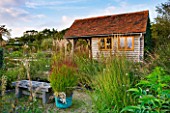 MARCHANTS HARDY PLANTS, EAST SUSSEX: GRAVEL GARDEN AND NURSERY SHED WITH COPPER CONTAINER PLANTED WITH GRASS - PANICUM VIRGATUM SHENANDOAH. GRAVEL, BENCH, GRASSES, COUNTRY