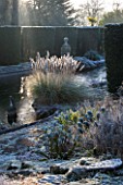 SEDGWICK PARK, WEST SUSSEX. COLUMNS OF YEW BESIDE THE LONG CANAL ALSO KNOWN AS THE WHITE SEA WITH PAMPAS GRASS AND FROSTED EUPHORBIAS. WINTER, GARDEN, FROST, JANUARY, WATER.