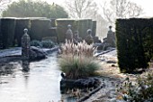 SEDGWICK PARK, WEST SUSSEX. COLUMNS OF YEW BESIDE THE LONG CANAL ALSO KNOWN AS THE WHITE SEA WITH PAMPAS GRASS. WINTER, GARDEN, FROST, JANUARY, WATER.